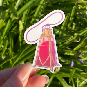 Peut inclure: Une bouteille en verre dorée et rouge avec un bouchon et un cordon rose. La bouteille est remplie d'un liquide rouge et présente un design décoratif.