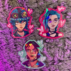 Puede incluir: Tres pegatinas holográficas con personajes de estilo anime con corazones rosas alrededor. Los personajes son una chica con cabello azul y un atuendo negro, una chica con cabello azul y un atuendo verde y azul, y una chica con cabello marrón y un atuendo negro.