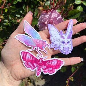 May include: Three colorful insect stickers on a hand. A pink and purple cicada, a pink and white moth, and a white and purple moth.