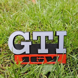 Puede incluir: Un emblema GTI 16V negro, rojo y plateado, sobre hierba verde. El emblema presenta las letras "GTI" en plata, sobre el texto rojo "16V". Las letras están delineadas en negro.