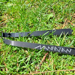 Könnte beinhalten: Zwei schwarze, rechteckige Objekte mit dem Wort "MANHATTAN" in weißen Buchstaben liegen auf grünem Gras. Die Objekte scheinen zerbrochen oder beschädigt zu sein. Das Gras ist üppig und grün, mit einigen braunen Flecken.