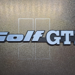 Op de afbeelding: Een zilver en wit "Golf GTI" embleem. De letters zijn vetgedrukt, zonder schreef, met een zilveren omtrek en witte vulling. Het embleem staat tegen een getextureerde, donkere achtergrond.