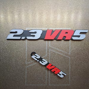 May include: Two automotive emblems featuring the text 2.3 VR5. The larger emblem has silver numbers and letters with a red VR section on a black background. The smaller emblem is a keychain version with the same silver, red, and black design.