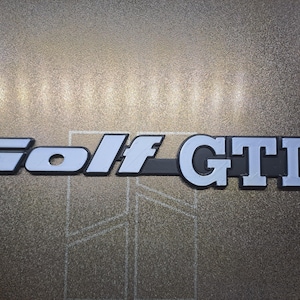 Puede incluir: Un emblema metálico plateado y negro con el texto Golf GTD en una fuente sans-serif en negrita. Las letras tienen un aspecto tridimensional en relieve con un acabado liso, sobre una superficie de fondo metálica oscura y texturizada.