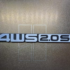 May include: A rectangular badge featuring the text "4WS 2.0 Si" in light grey. The text is set against a black background. The badge is displayed on a textured, metallic surface.
