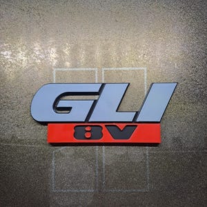 Puede incluir: Un emblema GLI 8V gris y negro con una base roja. Las letras GLI son de color gris claro con un contorno negro. El 8V es negro. El emblema está sobre una superficie oscura texturizada.