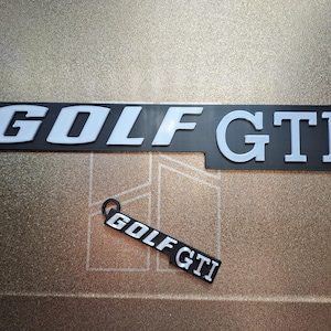 May include: A black rectangular emblem featuring the white text GOLF GTI in a bold, sans-serif font, accompanied by a smaller, matching black and white keychain tag that also displays the text GOLF GTI with a small circular ring attached.