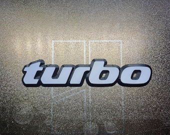 Może przedstawiać: Biały emblemat "turbo" z czarnym obramowaniem na teksturowanej szarej powierzchni.
