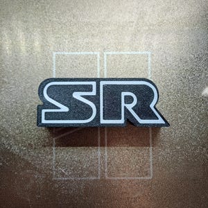 Puede incluir: Un logotipo en blanco y negro con las letras "SR" en una fuente en negrita y de bloque.