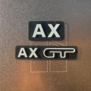 May include: Two black rectangular badges with white lettering. The top badge says "AX" and the bottom badge says "AX GT".