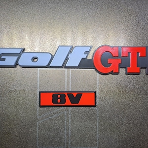 Può includere: Un set di emblemi per auto, tra cui un grande logo "Golf GTI" in grigio e rosso, e un emblema rettangolare più piccolo con "8V" in nero su sfondo rosso. Gli emblemi sono probabilmente per una Volkswagen Golf GTI.