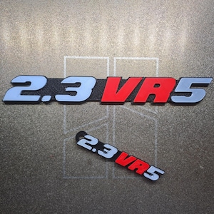 Peut inclure: Deux emblèmes automobiles avec le texte "2.3 VR5". Le plus grand emblème a le texte en bleu clair, rouge et blanc. Le plus petit emblème a le même texte dans les mêmes couleurs. Le fond est une surface grise texturée.