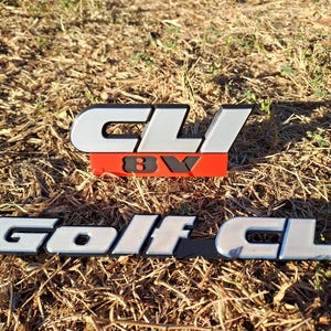 May include: Two automotive emblems featuring silver lettering with black outlines. The top emblem displays the text CLI above a red rectangular base with the black text 8V. The bottom emblem displays the text Golf CLI in a silver font on a black base.