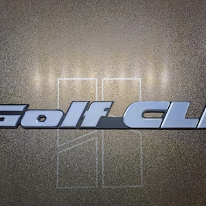 May include: A metallic emblem featuring the text Golf CLI in a bold, sans-serif font. The letters have a light gray, reflective finish with dark outlines. Behind the text, two vertical rectangular outlines are visible on a textured surface.