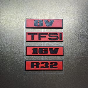 Puede incluir: Cuatro emblemas rectangulares para automóviles con texto negro sobre fondo rojo. Los emblemas dicen "8V", "TFSI", "16V" y "R32". Los emblemas están dispuestos verticalmente.