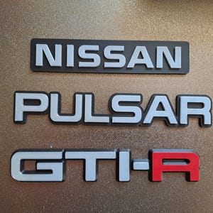 Puede incluir: Emblemas automotrices tridimensionales que presentan el texto NISSAN en plata sobre fondo negro, PULSAR en plata sobre fondo negro y GTI-R con letras plateadas y una R roja sobre fondo negro, dispuestos en tres filas.