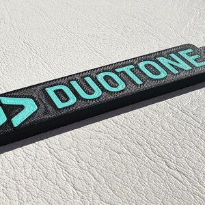 Peut inclure: Un porte-cl&eacute;s noir avec un logo bleu turquoise et le mot "DUOTONE" imprim&eacute; dessus.