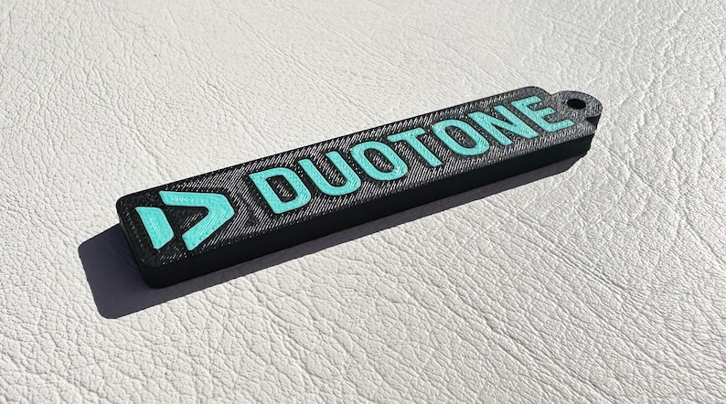 Peut inclure: Un porte-cl&eacute;s noir avec un logo bleu turquoise et le mot "DUOTONE" imprim&eacute; dessus.