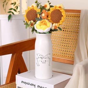 May include: A white ceramic vase with a bouquet of crocheted sunflowers, roses, and other flowers. The vase has a black line drawing of two elephants and the date 03.20.23. The flowers are yellow, orange, and white.