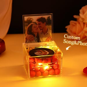 May include: A clear acrylic music box with a photo of a couple and a small record. The box is illuminated with warm white lights and filled with red decorative spheres. The text "Custom Song & Photo" is visible.