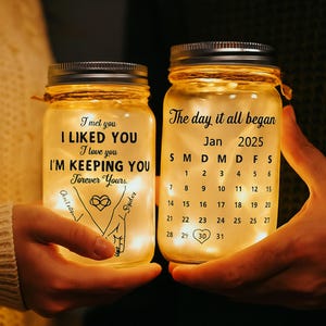 May include: Two illuminated mason jars with silver lids. One jar reads "I met you, I liked you, I love you, I'm keeping you, forever yours." The other jar displays a calendar for January 2025. Both jars are filled with fairy lights.