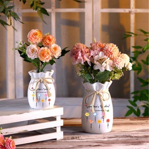 May include: Two white ceramic vases with a rope design and the text "Grandma's Garden" and a floral design. The vases are filled with pink and orange artificial flowers.
