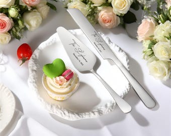 Puede incluir: Un juego de cuchillo y servidor de pastel de acero inoxidable con texto grabado. El juego está junto a un plato blanco con una magdalena cubierta con un corazón verde y un macaron rosa. El fondo incluye rosas blancas y rosas.