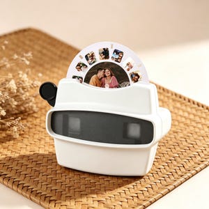 May include: A white retro-style viewer with a black eyepiece and a circular photo reel. The reel displays several small photos, including a couple. The viewer sits on a woven, brown surface.