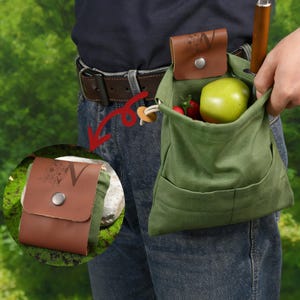 May include: A green canvas foraging bag with a brown leather pouch attached to a belt. The bag contains a green apple and red berries. A knife is visible. The leather pouch features a floral design and the letter "N".