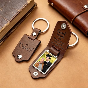 May include: Two brown leather keychains with silver key rings. One keychain features the word "Dad" with angel wings. The other keychain has a photo and the text "God Blessed The Broken Road That Let Me Straight to You."