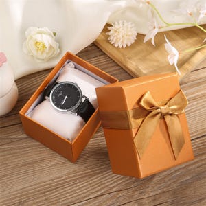 May include: A wristwatch with a black face and silver-tone case featuring a black leather strap, displayed inside an open orange gift box on a white cushion, with a matching orange box lid decorated with a gold ribbon bow placed beside it.