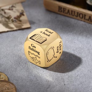 Pu&ograve; includere: Un dado di attivit&agrave; color oro con illustrazioni e testo neri. Le facce mostrano immagini e frasi come "Leggi un libro", "Vai a pescare" e "Fai un pisolino". Il dado &egrave; un regalo divertente.