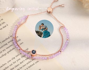 May include: A delicate rose gold bracelet with a purple and pink beaded chain. The bracelet has a sliding clasp and a small heart-shaped charm. The bracelet is displayed on a white surface with a book and a photo of a couple.