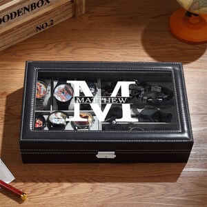 May include: Black leather watch box with a clear glass top. The box is divided into compartments holding watches, sunglasses, and a bracelet. The name "MATTHEW" is displayed in large white letters on the glass. The box is stitched with white thread.