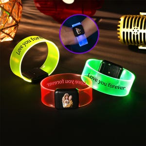 May include: Three illuminated wristbands in green, red, and yellow. Each band has a black rectangular component and the words "Love you forever" printed on them. One band displays a photo. A wrist is shown wearing a similar band.