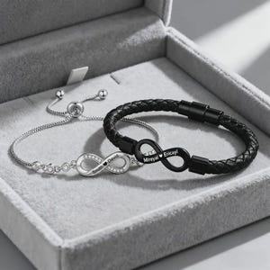Puede incluir: Dos pulseras en una caja gris. Una es plateada con un símbolo de infinito y los nombres "Smith" y "David". La otra es de cuero trenzado negro con un símbolo de infinito negro y los nombres "Mireyal" y "Ericayl".