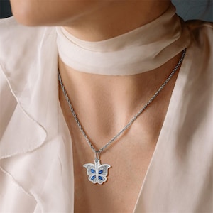 Puede incluir: Un collar de cadena de plata con un colgante de mariposa. La mariposa está decorada con piedras preciosas azules.