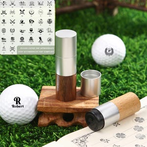 May include: A golf ball stamp set featuring a wooden and silver metal stamp, a small silver metal cup, and two golf balls. One golf ball has the letter "R" and the name "Robert" printed on it. The set is on a wooden stand.