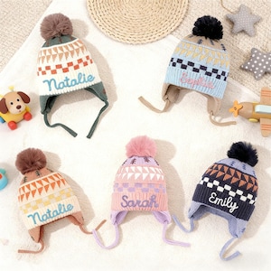 May include: A selection of children's winter hats with ear flaps and pom-poms. Each hat has a unique colour scheme and a name embroidered on the front. The hats feature geometric patterns and are designed for warmth and style.