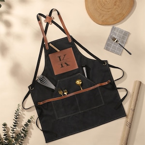 May include: Black apron with brown leather accents and straps. Features a front pocket with utensils and a brown leather patch with the name "Kevin" and the letter "K". Includes a whisk and a metal bottle.
