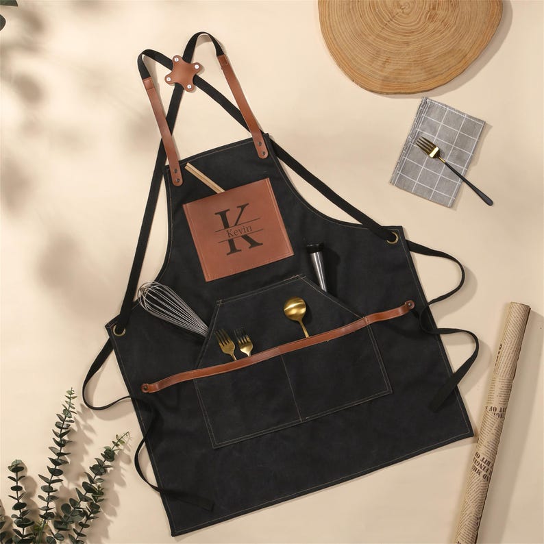 May include: Black apron with brown leather accents and straps. Features a front pocket with utensils and a brown leather patch with the name "Kevin" and the letter "K". Includes a whisk and a metal bottle.