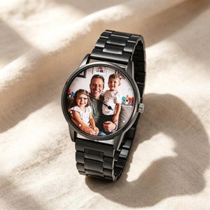 May include: A black metal wristwatch featuring a custom photo dial that displays a color image of an adult and two children. The watch has a round case, a link bracelet, and thin black hands. The dial includes small white circular hour markers.