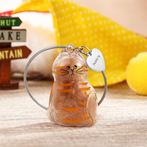 Puede incluir: Un llavero transparente con forma de gato con rayas y rasgos faciales naranjas. Un pequeño dije de corazón plateado con el nombre "Nancy" cuelga del llavero. El llavero está sujeto a un lazo de alambre plateado.