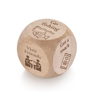 Pu&ograve; includere: Un dado di attivit&agrave; in legno con illustrazioni e testo incisi. Le facce mostrano immagini e frasi come "Vai a pescare", "Visita gli amici" e "Prendi uno spuntino". Il dado &egrave; marrone chiaro e poggia su una superficie bianca.