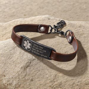 May include: A brown leather medical ID bracelet with a dark metal plate. The plate has a medical symbol and engraved text: Tammy Chase, ICE Sylvi 504-1108, Steve 380-4917, No BP/draw right arm.