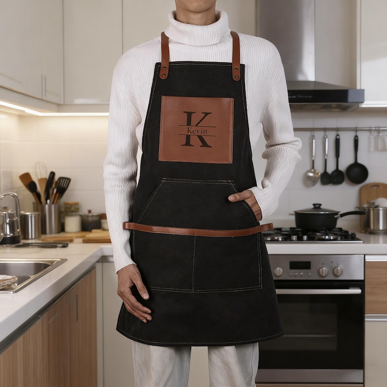 May include: Black apron with brown leather straps and accents. The apron features a large front pocket and a brown leather patch with the name "Kevin" and the initials "K" and "V". The apron is designed for kitchen or grilling use.