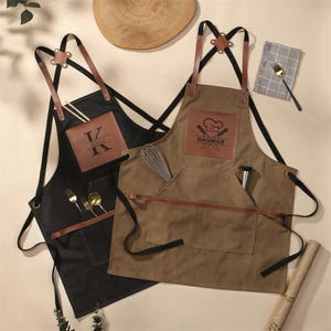 Puede incluir: Dos delantales, uno negro y otro marrón, con detalles de cuero y bolsillos. El delantal negro tiene las iniciales "K" y "V" en un parche de cuero. El delantal marrón presenta el nombre "DAGMAR" y un logotipo de gorro de chef. Ambos tienen utensilios.