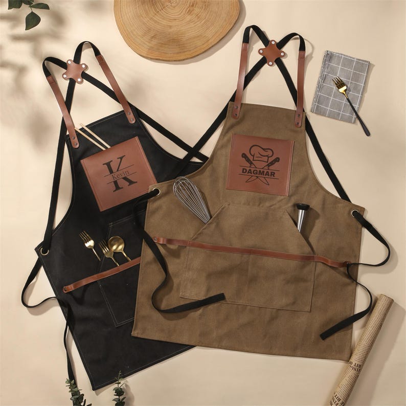 May include: Two aprons, one black and one brown, with leather accents and pockets. The black apron has the initials "K" and "V" on a leather patch. The brown apron features the name "DAGMAR" and a chef's hat logo. Both have utensils.