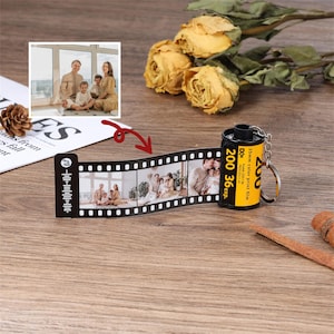 May include: A personalized film roll keychain with family photos. The black film strip displays multiple images, connected to a yellow and black film canister. The canister reads "200 36exp." and "35mm color print film".