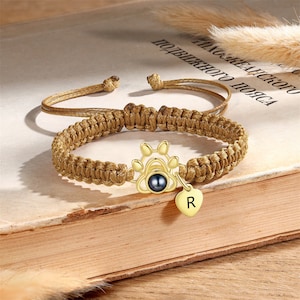 Puede incluir: Un brazalete trenzado marrón con un dije de pata de oro y un pequeño dije de corazón de oro con la letra "R" grabada. El brazalete está sobre un libro con una cubierta marrón.